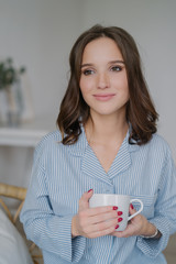 Indoor shot of dreamy young European woman with charming look, dressed in striped pyjamas, drinks coffee, thinks about something, enjoys calm domestic atmosphere. People, drinking and home concept