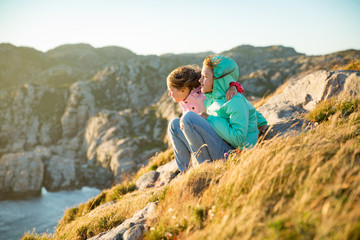 Two little girls play on rocky northern seashore. Sit, laugh, hug, explore the coastal rocks. Travel and enjoy a great adventure in Norway. Beautiful view of fjord and mountains in sunset.