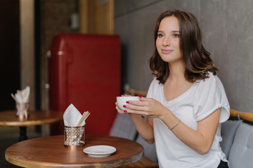 Pretty carefree young woman in casual white t shirt, has happy face, drinks aromatic coffee, spends free time in restaurant, sits at round wooden table, has thoughtful expression into distance.