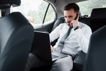 A young businessman working on laptop and talking on the phone while sitting in the car's back. Works in motion, appreciates its time