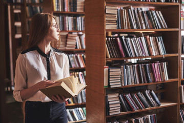 Young attractive student librarian reading a book between library bookshelves - obrazy, fototapety, plakaty
