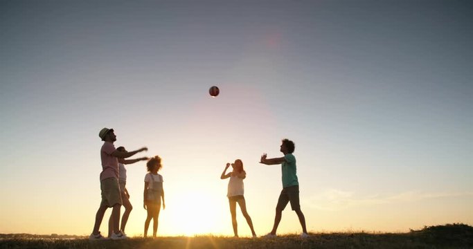 Silhouettes of active friends playing volleyball on a meadow near lake at sunset, and cheering. Friends having fun 4k