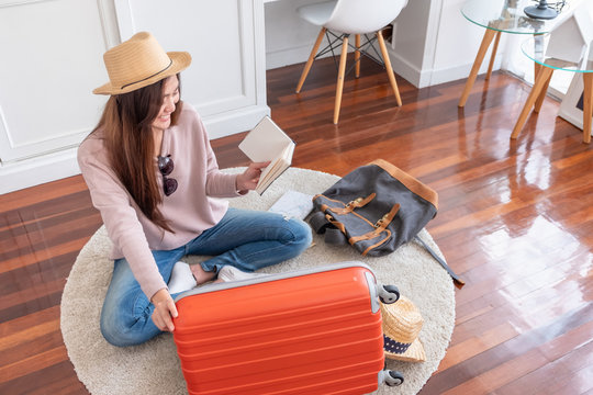 Young asian woman traveler prepare things into orange suitcase for holiday vacation at home.backpacker travel concept.