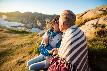 Loving mature couple hiking, sitting on windy top of rock, exploring. Active Mature man and woman wrapped in blanket, hugging and Happily smiling. Scenic view of sea, mountains. Norway, Lindesnes.