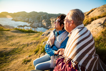 Loving mature couple hiking, sitting on windy top of rock, exploring. Active Mature man and woman wrapped in blanket, hugging and Happily smiling. Scenic view of sea, mountains. Norway, Lindesnes.