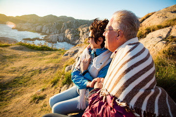 Loving mature couple hiking, sitting on windy top of rock, exploring. Active Mature man and woman wrapped in blanket, hugging and Happily smiling. Scenic view of sea, mountains. Norway, Lindesnes.