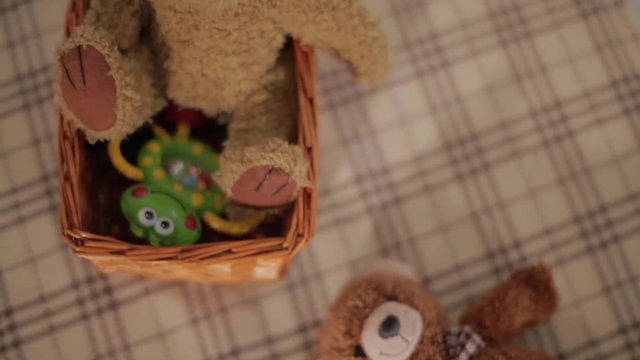 Smiling fluffy cute teddy bear is lying on its back on light brown checkered blanket next to small basket in room with dimmed light.