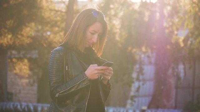 Attractive young girl stands in middle of busy buzzling city on summer sunset. Gorgeous young woman in a bright sunlight uses her phone.