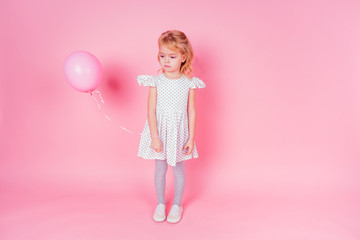 Obraz na płótnie Canvas sad little blonde girl in white dress in peas 4-5 year old holding balloon in the studio on a pink background,birthday celebration ,sadness and disappointment sorrow
