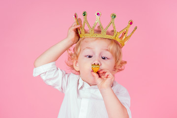 Obraz na płótnie Canvas little blond boy with golden crown head curls hairstyle 4-5 year old in studio on pink background blowing noisemakers horn-whistle a birthday party celebrates Christmas and New Year king of the party