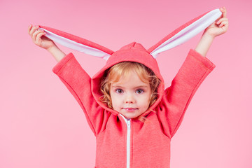 beautiful charming little girl curly blonde hairstyle wearing red sweater clothes with hood rabbit ears.Adorable child celebrate Easter,playing theater role of the easter bunny pink background studio