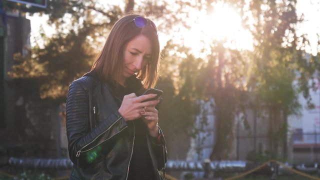 Attractive young girl stands in middle of busy buzzling city on summer sunset. Gorgeous young woman in a bright sunlight uses her phone.