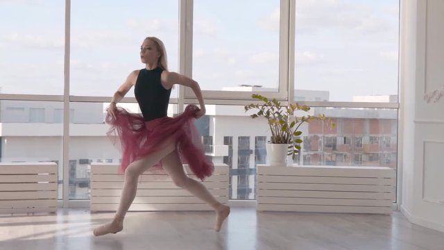 professional ballet dancer jumping into the splits against the backdrop of a high window. woman in a black bathing suit burgundy tutu on pointe. the dancer runs up makes an elegant light jump
