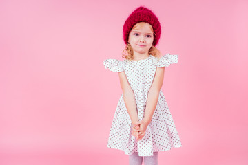portrait of happy charming little girl wearing knitted red hat and cute white dress on pink background in studio. Fashion autumn winter season sale concept.making a wish christmas night and birthday