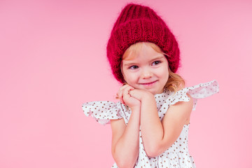 portrait of happy charming little girl wearing knitted red hat and cute white dress on pink background in studio. Fashion autumn winter season sale concept.making a wish christmas night and birthday