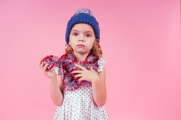 Beautiful little girl in dress and blue knitted hat in studio on white background. Happy little smiling female child with shiny Christmas tinsel. Christmas miracles Happy New Year