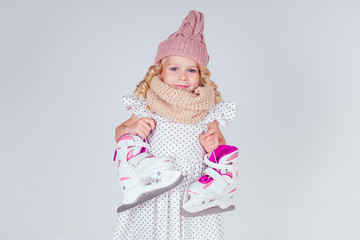 beautiful curly blonde little girl in warm knitted beige hat holding sportive figure skates white background in a studio. active life of a sports child