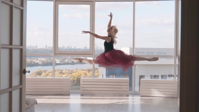 professional ballet dancer jumping into the splits against the backdrop of a high window. woman in a black bathing suit burgundy tutu on pointe. the dancer runs up makes an elegant light jump