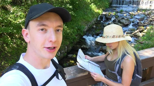 A young attractive hiking couple in a forest, the man talks to the camera, the woman reads a map - a creek in the background