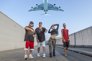 group of rappers posing on the metal rooftops of an abandoned building while a plane flies overhead