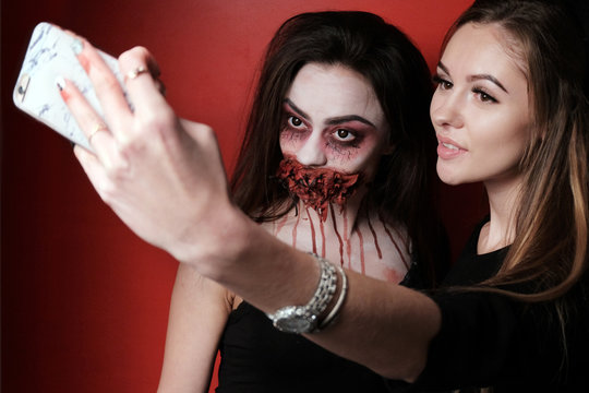 Concept selfie makeup for the story in the style of Halloween. A beautiful girl together with another girl in the image of a witch are taking pictures of themselves on the phone. Red background.