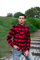 Naklejka na ściany i meble Portrait of stylish male student with bread posing outdoors, looking at camera. Handsome man wearing in red checked shirt walking in autumn park. Concept of study, and mans fashion.