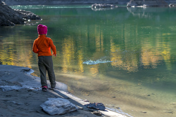 A little girl, a child, in warm clothes and a red cap throws stones into the water from the sandy shore