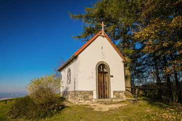 Obraz premium Autumn landscape, view of white chapel of Saint Mary Magdalene with red roof, wooden cross on top of front wall, iron bars on door, bright sunny day, clear blue sky, trees, grass, dry leaves on ground