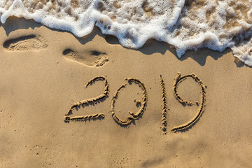 Concept of Happy New Year 2019 is coming and leaving year of 2018. Text on the sea beach and footprints on the sand. Wave water covering digits inscription. Meet at a tropical resort in Thailand.