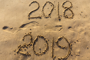 Concept of Happy New Year 2019 is coming and leaving year of 2018. Text on the sea beach and footprints on the sand. Wave water covering digits inscription. Meet at a tropical resort in Thailand.