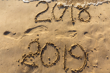 Concept of Happy New Year 2019 is coming and leaving year of 2018. Text on the sea beach and footprints on the sand. Wave water covering digits inscription. Meet at a tropical resort in Thailand.