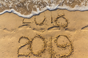 Concept of Happy New Year 2019 is coming and leaving year of 2018. Text on the sea beach and footprints on the sand. Wave water covering digits inscription. Meet at a tropical resort in Thailand.
