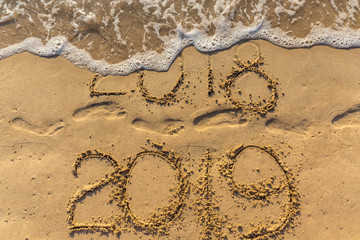 Concept of Happy New Year 2019 is coming and leaving year of 2018. Text on the sea beach and footprints on the sand. Wave water covering digits inscription. Meet at a tropical resort in Thailand.