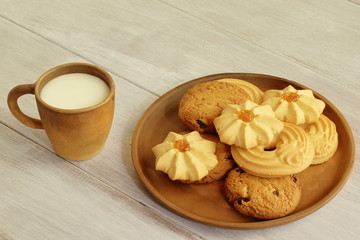 Tasty breakfast. Hot milk in clay cup and fragrant crumbly cookies with raisins nuts and jam on ceramic plate on light brown wooden countertop