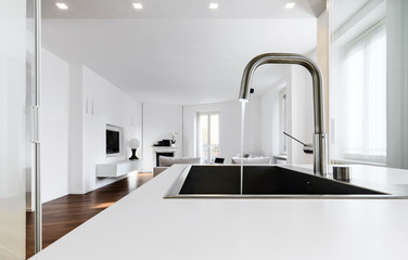 modern kitchen interiors in the foreground the integrated steel sink and the chrome faucet that overlooking on the living area - obrazy, fototapety, plakaty