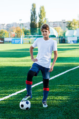 Obraz premium cute young boy in white blue sportswear stands next to classical black and white football ball on the stadium field. Soccer game, training, hobby concept. 
