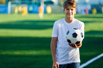 cute young boy in white blue sportswear holds classical black and white football ball on the stadium field. Soccer game, training, hobby concept. 