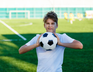 cute young boy in white blue sportswear holds classical black and white football ball on the stadium field. Soccer game, training, hobby concept. 