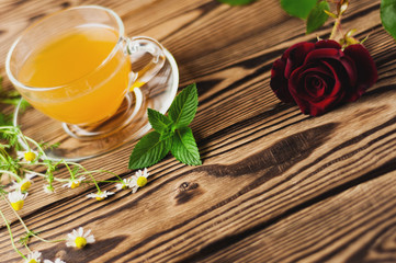 Glass cup with green tea and mint and bunch of chamomile near red rose on sprig on old wooden...