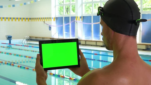 A swimmer looks at a tablet with a green screen at an indoor pool - closeup from behind
