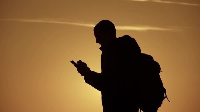 A silhouette young tourist man with a backpack uses a mobile phone. In the rays of the setting sun. Slow motion. Travel concept. Always online.