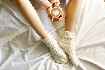 Young woman drinking cappuccino coffee and sitting in bed with white linnen. Top view of female legs in warm wool socks & sweater. Comfort winter holidays concept. Close up, copy space, background.