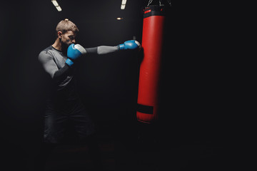 Young male boxer hitting punching bag on black background in gym.