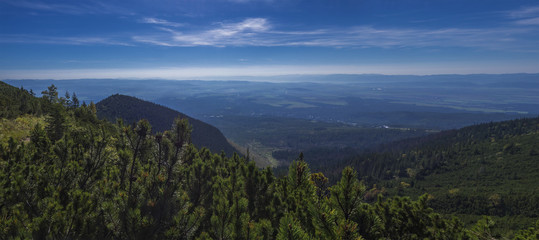 Panoramic view from tatra mountains trail on valley with Tatranska lomnice and blue misty slopes of hills in the distance. Pine trees and coniferous forest hills, blue sky. Tatra mountain in summer.