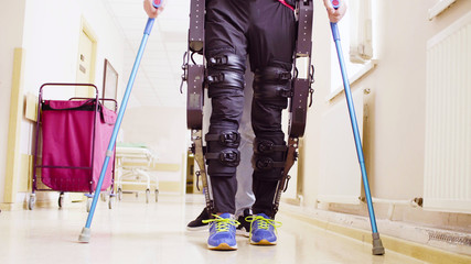 Legs of disable man in the robotic exoskeleton walking through the corridor of the rehabilitation clinic. Doctor helping him. - obrazy, fototapety, plakaty