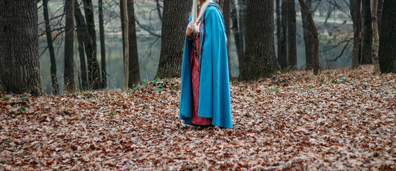 a girl with long hair, blonde, in a blue cape and a pink dress, walks through the autumn forest, along the yellow foliage of fallen leaves. A girl's sheath hangs on her belt.