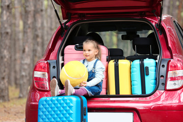 Cute little girl sitting in car trunk loaded with suitcases on forest road