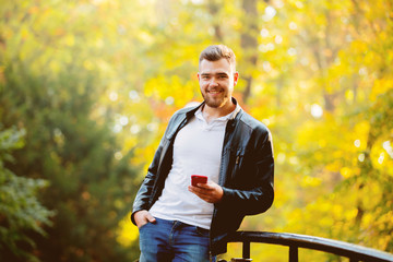 Obraz premium Young white guy using a mobile phone in a park with yellow trees on background. Autumn season time