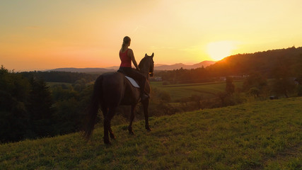 LENS FLARE Unrecognizable young woman looks at the sunset while sitting on horse