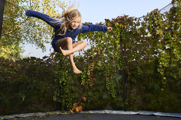 Child cute blond girl playing and jumping on trampoline with greenery background